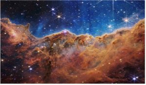 Carina Nebula image taken by the JWST. The image is divided horizontally by a curved line like a mountain range forming orange-brown clouds at the bottom of the image. The top of image is blue with bright stars of different sizes speckled across.