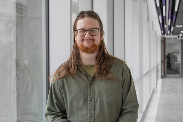 A photo of David Fortin, a man with long brown hair and glasses standing in the hallway.
