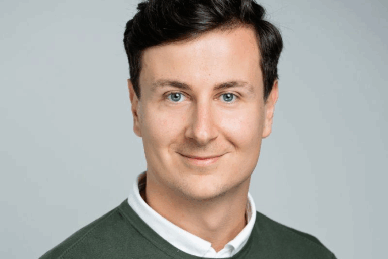 A photo of Dr. Marko Biberdzic, smiling at the camera in a collared white shirt with a green sweater over it. He's against a blank white background.