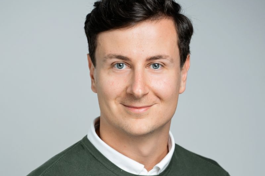 A photo of Dr. Marko Biberdzic, smiling at the camera in a collared white shirt with a green sweater over it. He's against a blank white background.