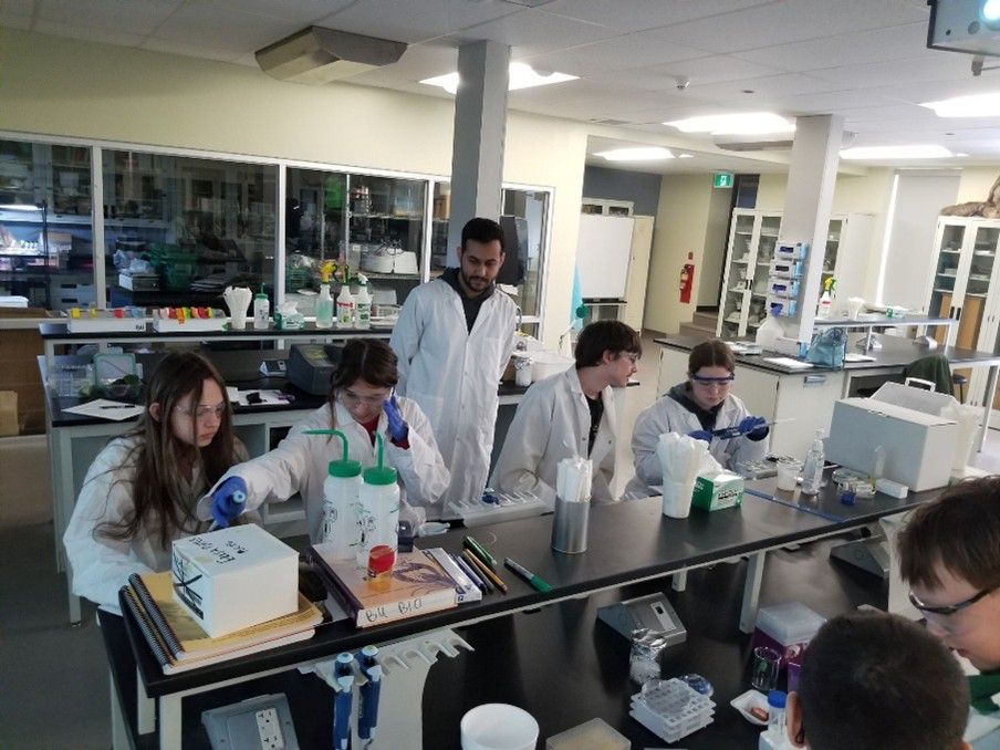 A group of young scientists in full technician gear (lab coats, goggles, gloves) work together in a lab with an instructor looking over them.