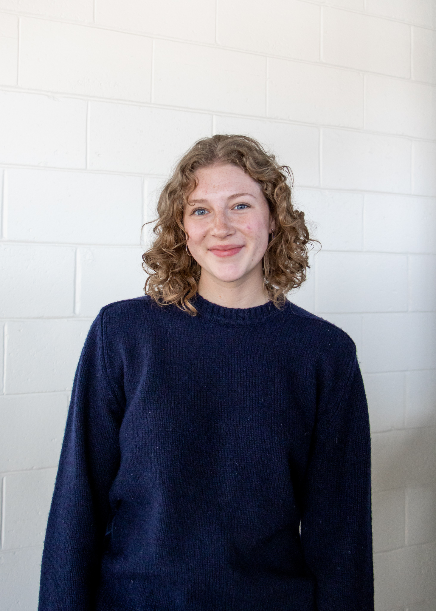 A young woman with light brown curly hair is in front of a white wall. She is Grace Johnston.