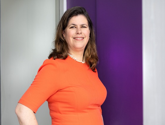 Picture of Dr. Heather Lawford in an orange dress in front of a purple and grey wall. She's smiling.