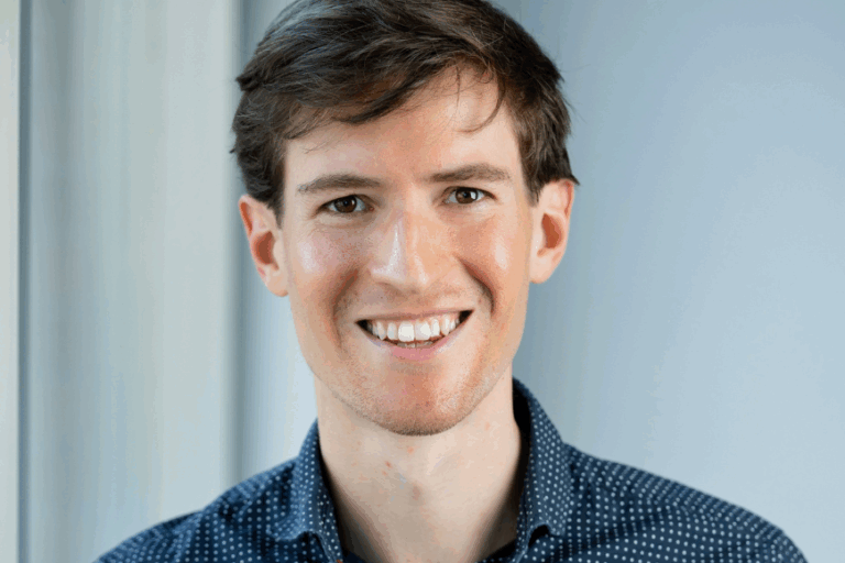 A picture of Dr. Jacob Robbins-Kanter. She's in a blue dotted shirt, has brown hair, and smiles at the camera in a portrait shot set in a white hallway.