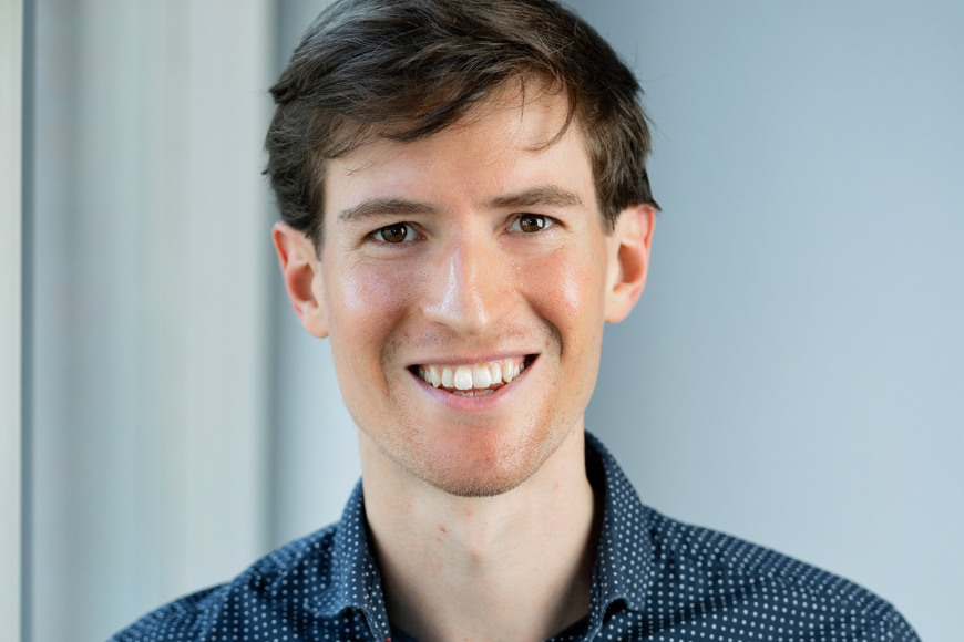 A picture of Dr. Jacob Robbins-Kanter. She's in a blue dotted shirt, has brown hair, and smiles at the camera in a portrait shot set in a white hallway.
