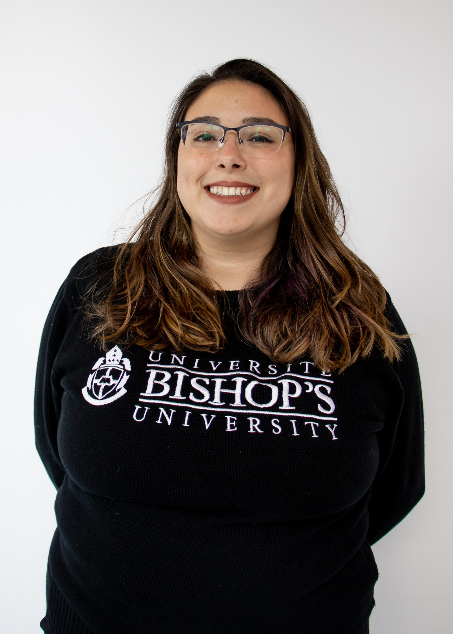 A photo of a young woman in a Bishop's hoodie, glasses, and brown hair in front of a white wall. She is Maria Carolina Lopez.