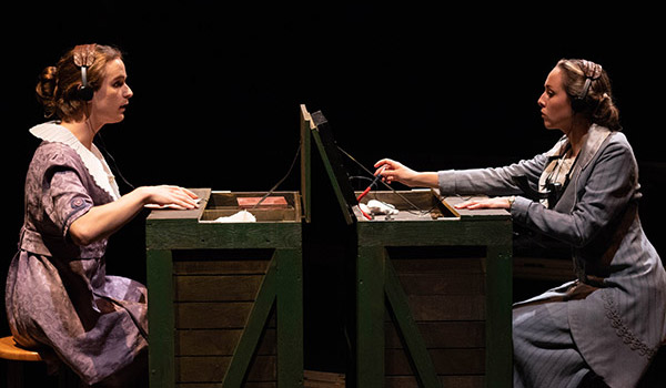 Two students seated at vintage telephone switchboards, wearing period attire, focused on their work for one of the Bishop's play.