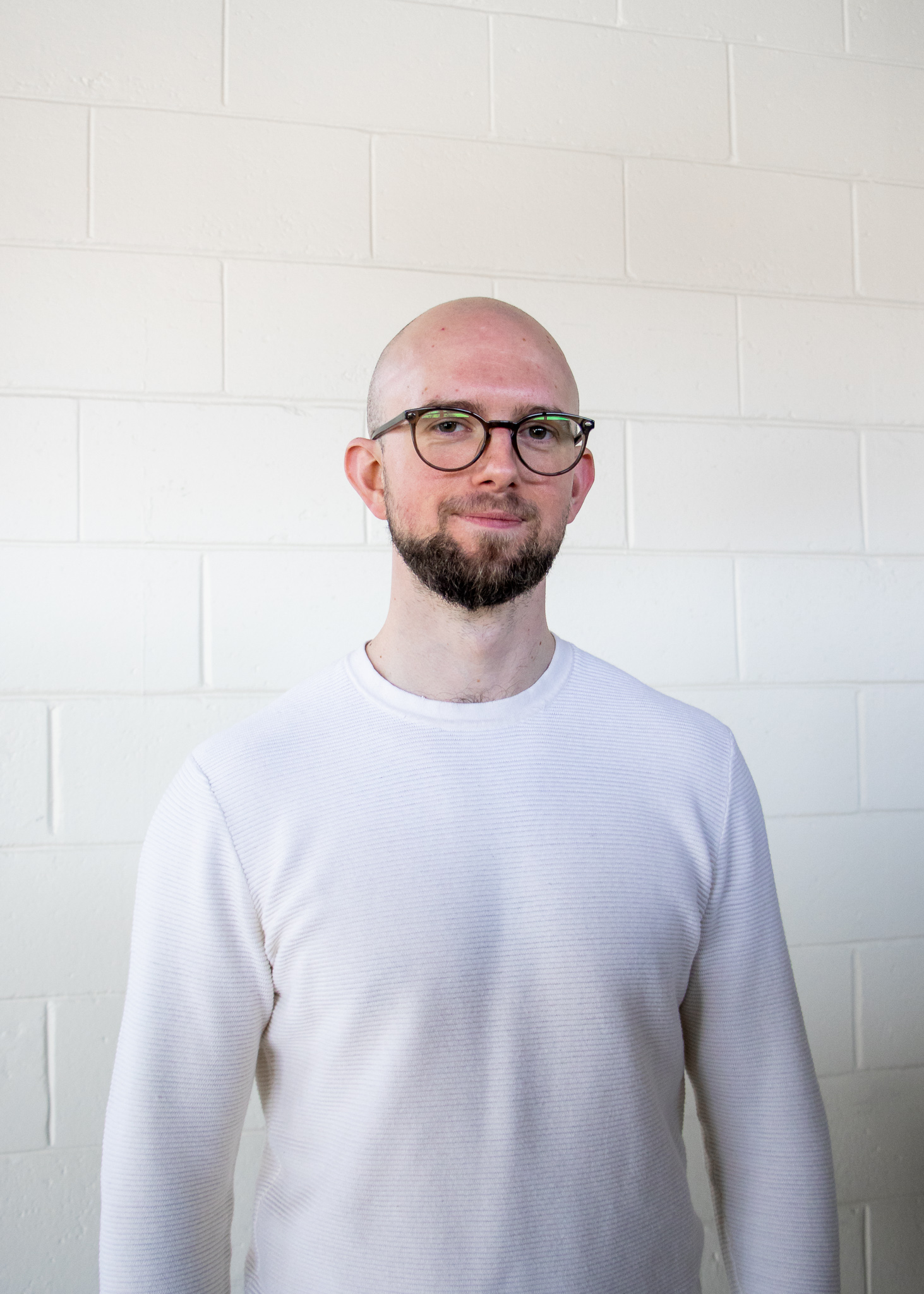 A young man with glasses and a shaved head stands in front of a white wall. He is Zachary Fry.