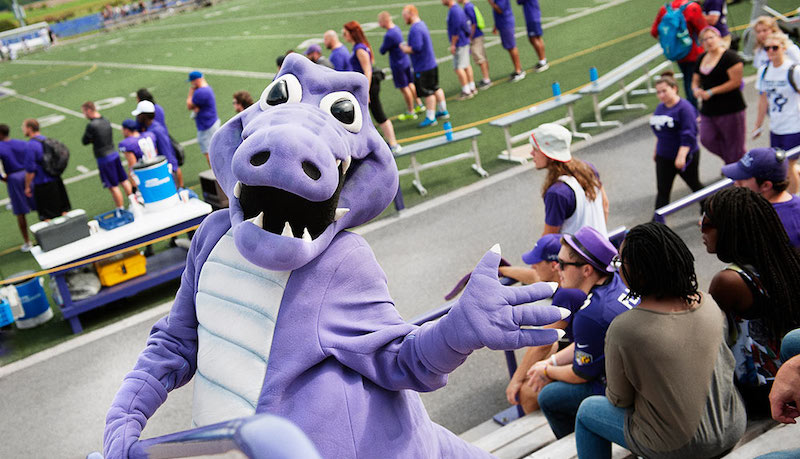 The Bishop's Gaiter (a large, purple alligator mascot, with gaiters on its legs) smiles and waves to the camera during a home football game.